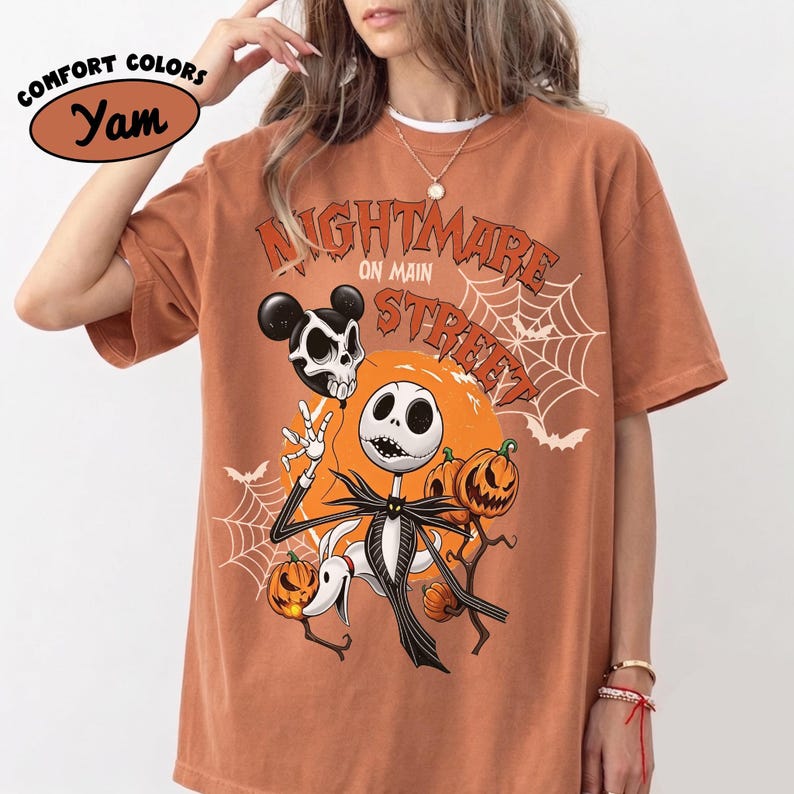 Pu&ograve; includere: Una t-shirt a maniche corte color arancio bruciato con una stampa grafica recante il testo "NIGHTMARE ON MAIN STREET". Il design include un personaggio scheletrico, un cane fantasma, zucche, ragnatele, pipistrelli e un palloncino a forma di teschio su una luna arancione.