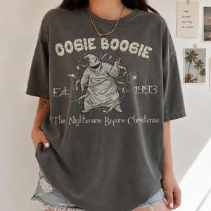 May include: A charcoal grey short-sleeve t-shirt featuring a graphic of Oogie Boogie from The Nightmare Before Christmas. The print includes the text OOGIE BOOGIE, Est. 1993, and The Nightmare Before Christmas, along with spider web illustrations.
