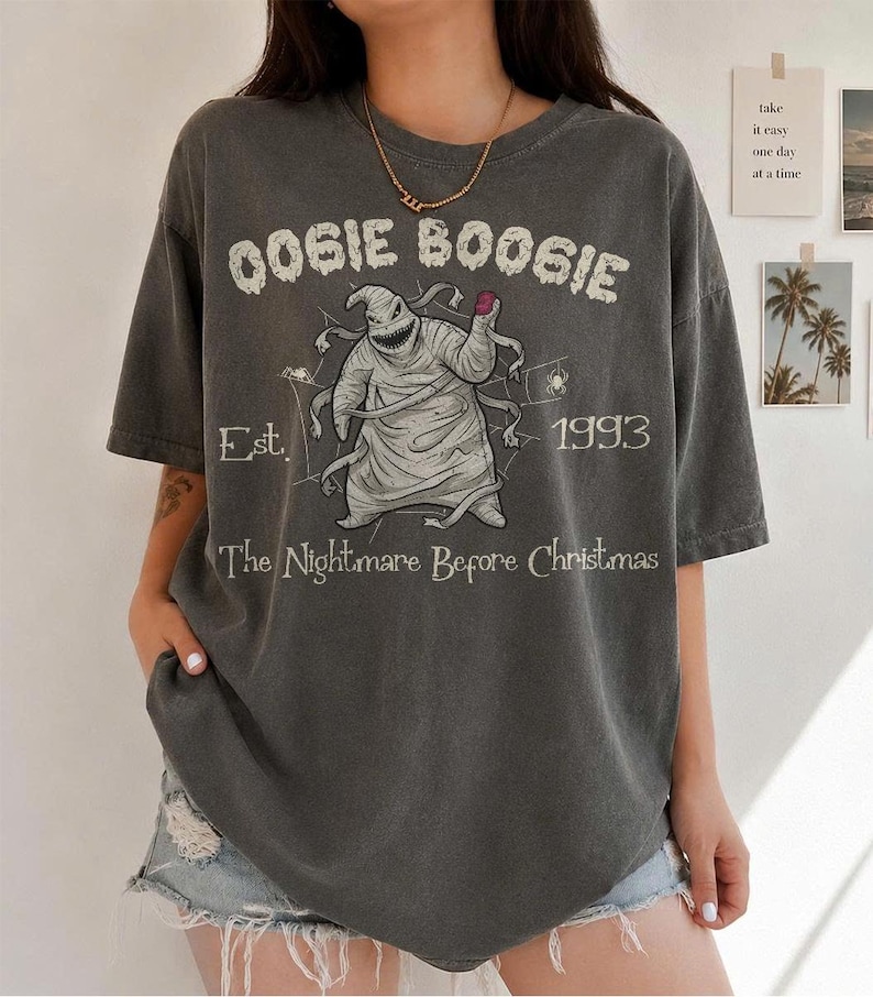 May include: A charcoal grey short-sleeve t-shirt featuring a graphic of Oogie Boogie from The Nightmare Before Christmas. The print includes the text OOGIE BOOGIE, Est. 1993, and The Nightmare Before Christmas, along with spider web illustrations.