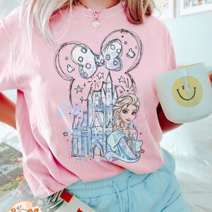 Puede incluir: Una camiseta rosa con un gr&aacute;fico de un castillo, un lazo y una princesa. La princesa tiene el pelo largo y rubio y lleva un vestido azul. El gr&aacute;fico est&aacute; en estilo de boceto y tiene un efecto acuarela.