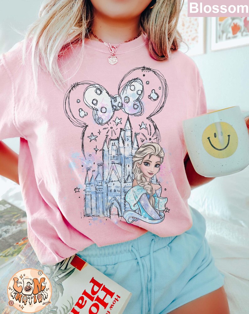 Puede incluir: Una camiseta rosa con un gr&aacute;fico de un castillo, un lazo y una princesa. La princesa tiene el pelo largo y rubio y lleva un vestido azul. El gr&aacute;fico est&aacute; en estilo de boceto y tiene un efecto acuarela.