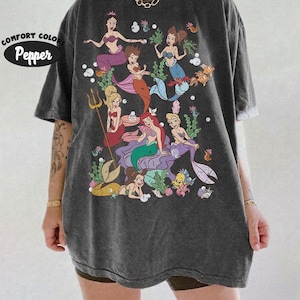 Puede incluir: Una camiseta de manga corta gris carb&oacute;n con un estampado gr&aacute;fico de varias sirenas en diversas poses con elementos acu&aacute;ticos como burbujas, algas y peces. El texto Comfort Colors Pepper est&aacute; impreso en la esquina superior izquierda de la prenda.