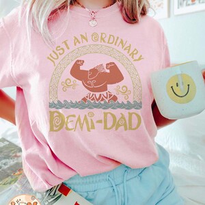 以下が含まれることがあります： 「Just an Ordinary Demi-Dad」というテキストと、漫画のキャラクターの金色のグラフィックが描かれたピンクのTシャツ。