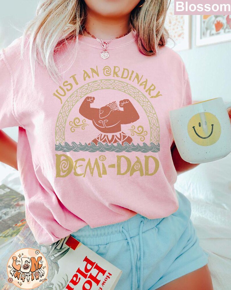 以下が含まれることがあります： 「Just an Ordinary Demi-Dad」というテキストと、漫画のキャラクターの金色のグラフィックが描かれたピンクのTシャツ。