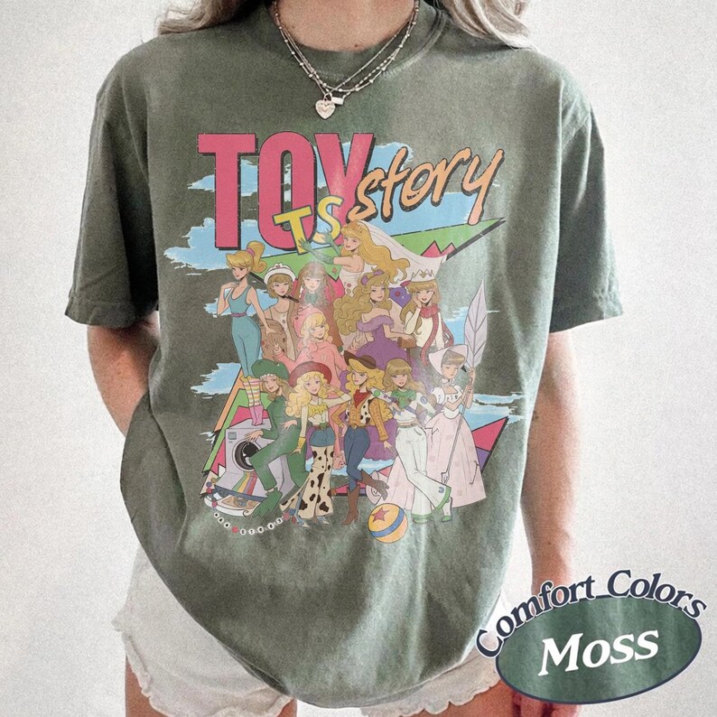 Peut inclure: Un t-shirt &agrave; manches courtes vert mousse avec une impression graphique portant le texte "TOY story" en rose et jaune. Le motif montre un groupe de personnages illustr&eacute;s dans diverses tenues, incluant un ballon, sur un fond de ciel bleu.