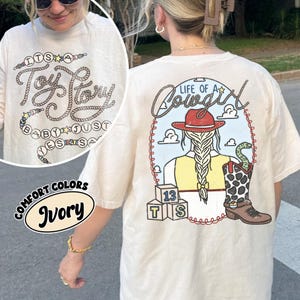 May include: An ivory t-shirt featuring a graphic on the back of a cowgirl with a braided ponytail, a red hat, and a cow-print boot with a snake. The text reads Life of a Cowgirl, with blocks labeled 13, T, and S. The front shows Toy Story text.