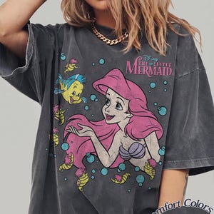 May include: A charcoal gray short-sleeve t-shirt featuring a graphic print of Ariel from The Little Mermaid with Flounder and seahorses. The design includes the text Disney The Little Mermaid in pink and blue bubbles. The shirt has a faded, vintage look.