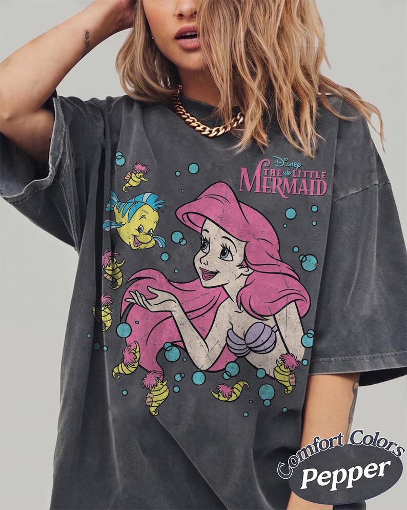 May include: A charcoal gray short-sleeve t-shirt featuring a graphic print of Ariel from The Little Mermaid with Flounder and seahorses. The design includes the text Disney The Little Mermaid in pink and blue bubbles. The shirt has a faded, vintage look.
