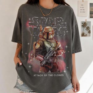 K&ouml;nnte beinhalten: Dunkelgraues T-Shirt mit einem Star Wars-Motiv von Boba Fett aus Angriff der Klonkrieger. Das Design beinhaltet das Star Wars-Logo und eine Darstellung der Figur in ihrer R&uuml;stung, die einen Blaster h&auml;lt. Das Shirt hat einen Vintage-Look.