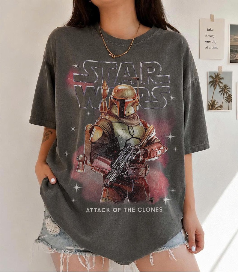 K&ouml;nnte beinhalten: Dunkelgraues T-Shirt mit einem Star Wars-Motiv von Boba Fett aus Angriff der Klonkrieger. Das Design beinhaltet das Star Wars-Logo und eine Darstellung der Figur in ihrer R&uuml;stung, die einen Blaster h&auml;lt. Das Shirt hat einen Vintage-Look.