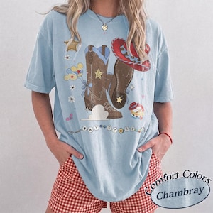 Pu&ograve; includere: Una t-shirt azzurro chiaro con una stampa grafica di stivali da cowboy marroni, un cappello da cowboy rosso, una stella da sceriffo, palloncini e fiori. Il testo TAY STORY &egrave; stampato in stile perline nella parte inferiore del disegno frontale.