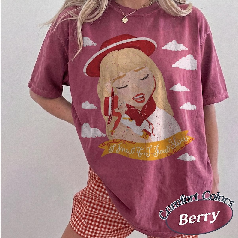 Pode incluir: Uma t-shirt de manga curta cor de baga com uma ilustra&ccedil;&atilde;o gr&aacute;fica de uma personagem loira com um chap&eacute;u e um cachecol vermelhos. O design inclui nuvens brancas e uma faixa amarela com o texto I Knew It, I Knew You. Gola redonda.