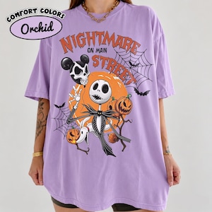 Pu&ograve; includere: Una t-shirt color orchidea con una stampa grafica recante il testo Nightmare on Main Street. Il design include un personaggio scheletro, un cane fantasma, zucche, ragnatele e pipistrelli su uno sfondo circolare arancione.