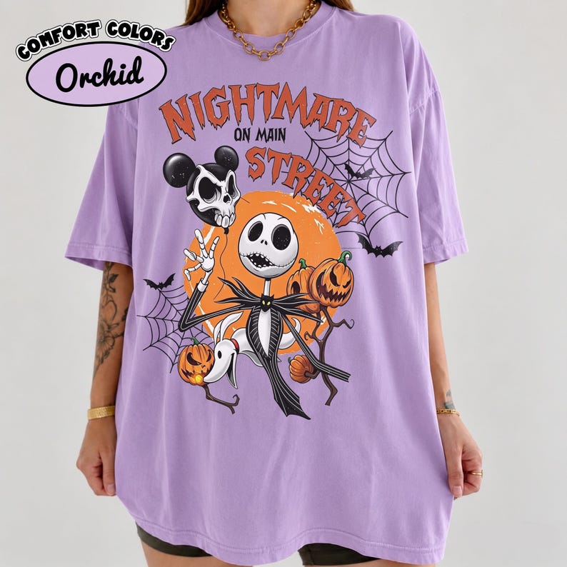 Pu&ograve; includere: Una t-shirt color orchidea con una stampa grafica recante il testo Nightmare on Main Street. Il design include un personaggio scheletro, un cane fantasma, zucche, ragnatele e pipistrelli su uno sfondo circolare arancione.