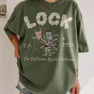 May include: An olive green t-shirt featuring the text LOCK in large white letters, a graphic of a mummy character holding a mask in front of a spider web, the text Est. 1993, and the text The Nightmare Before Christmas printed in white.