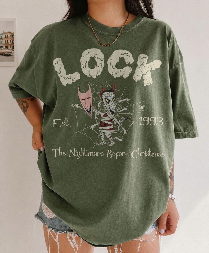 May include: An olive green t-shirt featuring the text LOCK in large white letters, a graphic of a mummy character holding a mask in front of a spider web, the text Est. 1993, and the text The Nightmare Before Christmas printed in white.