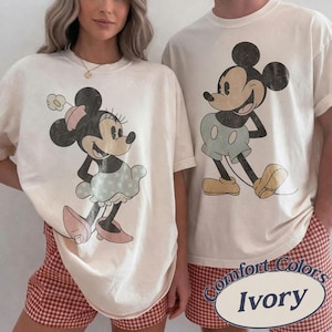 May include: Two ivory t-shirts featuring vintage-style graphic prints of Mickey and Minnie Mouse. The left shirt displays Minnie Mouse with a flower hat and polka dot skirt, while the right shirt shows Mickey Mouse in shorts and shoes.