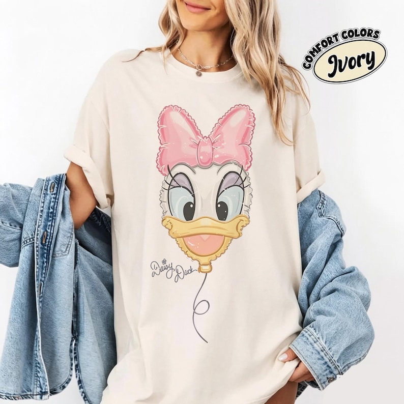 Pode incluir: Uma t-shirt cor de marfim com um gr&aacute;fico da Daisy Duck desenhado como um bal&atilde;o cor-de-rosa com um la&ccedil;o. O gr&aacute;fico inclui o texto Daisy Duck em letra cursiva. Um emblema circular no canto superior direito exibe o texto Comfort Colors Ivory.