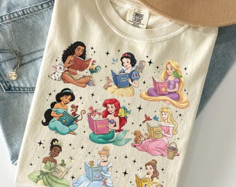 Puede incluir: Camiseta de color crema con ilustraciones de nueve princesas de Disney leyendo libros. Cada princesa está representada con su aspecto característico y rodeada de personajes y objetos relacionados. La camiseta está hecha de un material suave y cómodo.