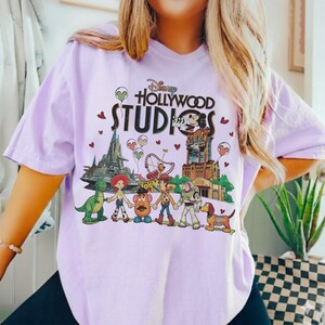 Op de afbeelding: Lila T-shirt met een kleurrijke afbeelding van Disney Hollywood Studios. Het ontwerp toont Toy Story-personages, een dinosaurus en het logo van het park. Het shirt heeft korte mouwen en een casual stijl, perfect voor dagelijks gebruik.