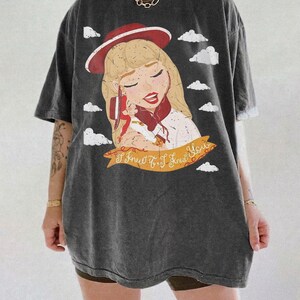 Pode incluir: Uma t-shirt cinzenta escura oversize com uma ilustra&ccedil;&atilde;o gr&aacute;fica de uma pessoa loira com um chap&eacute;u e um len&ccedil;o vermelhos, segurando uma boneca pequena. O design inclui nuvens brancas e uma faixa amarela com o texto I Knew It, I Knew You.