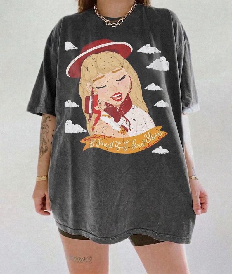 Pode incluir: Uma t-shirt cinzenta escura oversize com uma ilustra&ccedil;&atilde;o gr&aacute;fica de uma pessoa loira com um chap&eacute;u e um len&ccedil;o vermelhos, segurando uma boneca pequena. O design inclui nuvens brancas e uma faixa amarela com o texto I Knew It, I Knew You.