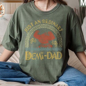 以下が含まれることがあります： 「Just an Ordinary Demi-Dad」というテキストと、漫画のキャラクターのグラフィックデザインが施された緑色のTシャツです。