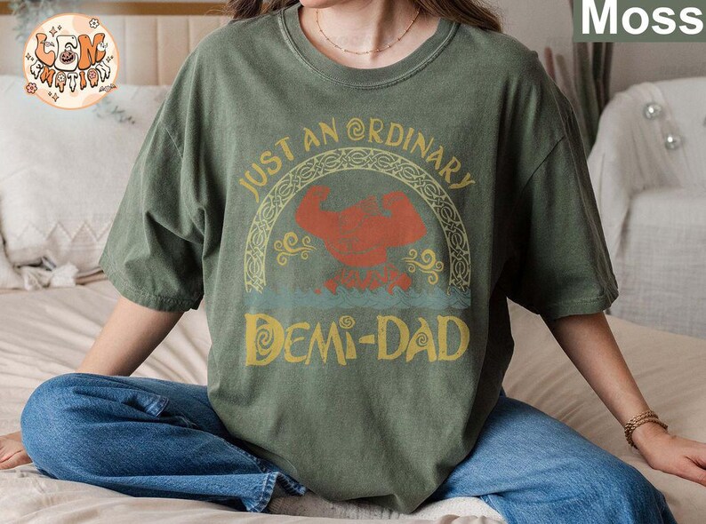 以下が含まれることがあります： 「Just an Ordinary Demi-Dad」というテキストと、漫画のキャラクターのグラフィックデザインが施された緑色のTシャツです。