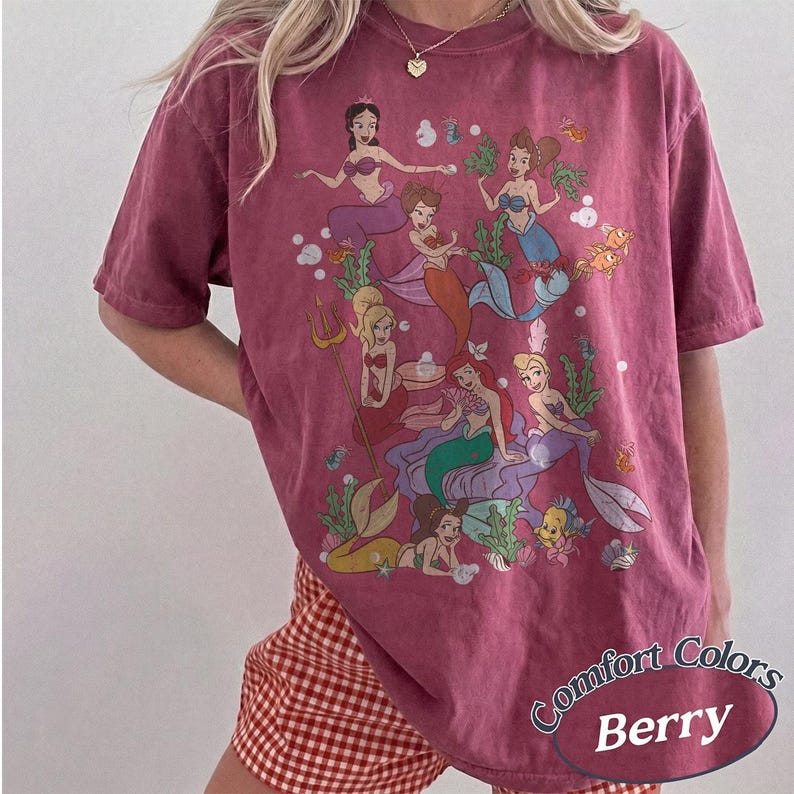 Puede incluir: Una camiseta de manga corta color baya con un estampado gr&aacute;fico de varias sirenas con diferentes colores de pelo y colas, rodeadas de elementos submarinos como algas, burbujas y peces peque&ntilde;os. El texto Comfort Colors Berry es visible.