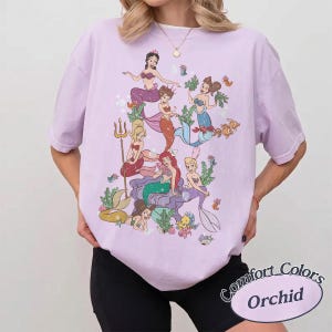 Puede incluir: Una camiseta de manga corta de color morado claro con un estampado gr&aacute;fico de varias sirenas de dibujos animados con diversos colores de pelo y dise&ntilde;os de cola, rodeadas de peque&ntilde;os animales marinos y plantas acu&aacute;ticas.