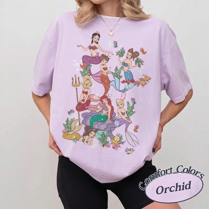 Puede incluir: Una camiseta de manga corta de color morado claro con un estampado gr&aacute;fico de varias sirenas de dibujos animados con diversos colores de pelo y dise&ntilde;os de cola, rodeadas de peque&ntilde;os animales marinos y plantas acu&aacute;ticas.