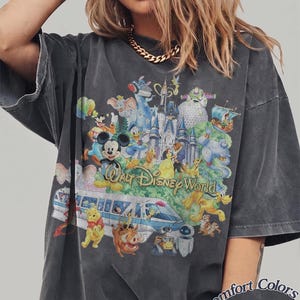 May include: A charcoal gray short-sleeve t-shirt featuring a large, colorful graphic print of various Disney characters, the Walt Disney World castle, and a monorail. The text "Walt Disney World" is printed in the center. "Comfort Colors Pepper" is visible.