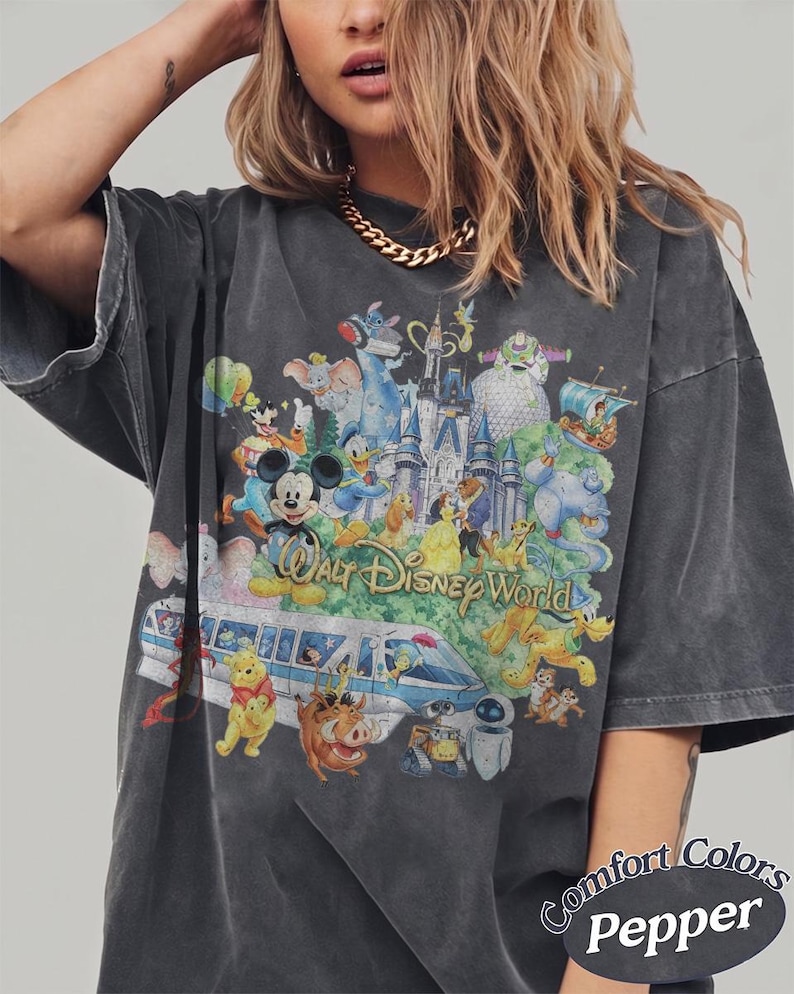 May include: A charcoal gray short-sleeve t-shirt featuring a large, colorful graphic print of various Disney characters, the Walt Disney World castle, and a monorail. The text "Walt Disney World" is printed in the center. "Comfort Colors Pepper" is visible.