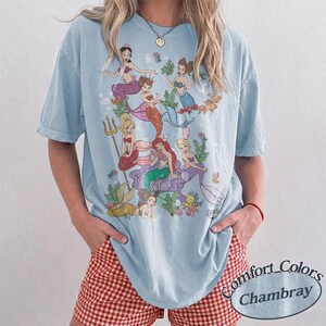 Puede incluir: Una camiseta de manga corta azul claro con un estampado gr&aacute;fico de varias sirenas con colas coloridas, algas y elementos acu&aacute;ticos. La camiseta tiene cuello redondo y un corte holgado. Una etiqueta peque&ntilde;a muestra el texto Comfort Colors Chambray.