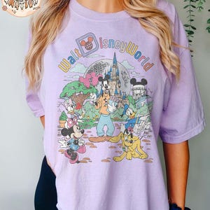 May include: A purple t-shirt with a vintage-style graphic of Mickey Mouse and friends at Walt Disney World. The text "Walt Disney World" is written in a colorful arc around the characters.