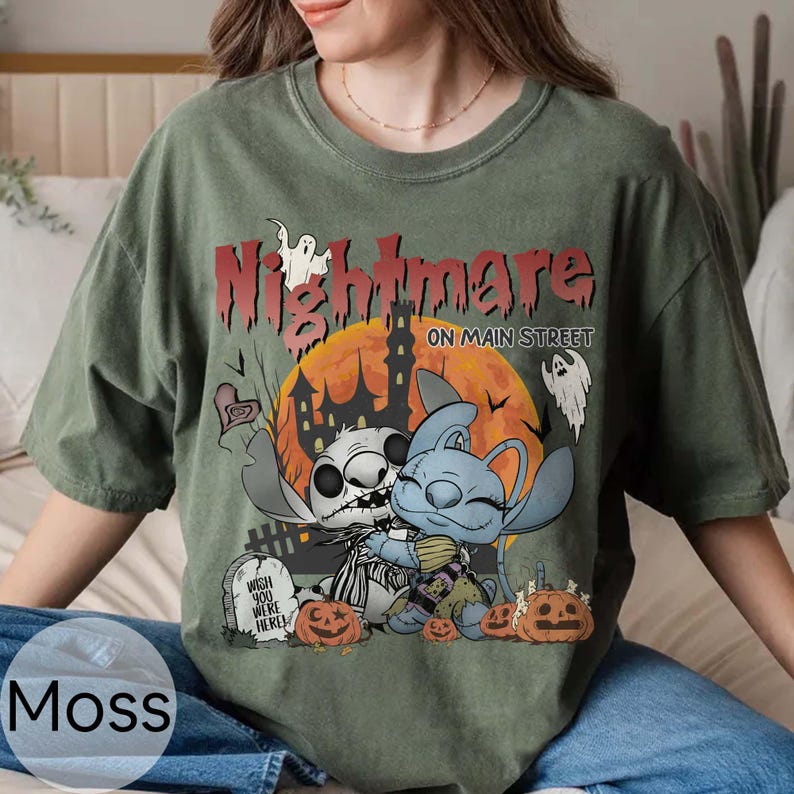Puede incluir: Camiseta verde musgo con un gr&aacute;fico tem&aacute;tico de Halloween. El dise&ntilde;o presenta dos personajes de dibujos animados abraz&aacute;ndose frente a una casa embrujada y una luna llena. El texto "Nightmare on Main Street" est&aacute; encima de los personajes.