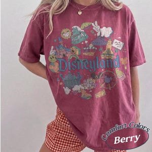 May include: Berry-colored t-shirt with a Disneyland graphic. The design features various park icons, including a castle, a Ferris wheel, and the Disneyland logo. The shirt has short sleeves and a relaxed fit. The words "Comfort Colors" and "Berry" are printed on the bottom.