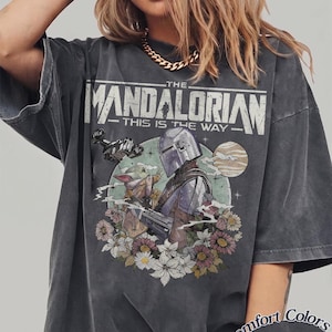 May include: A charcoal gray short-sleeve t-shirt featuring a graphic print of The Mandalorian and Grogu. The design includes the text THE MANDALORIAN THIS IS THE WAY, a spaceship, a planet, and colorful flowers. The shirt has a Comfort Colors Pepper label.