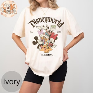 K&ouml;nnte beinhalten: Ein wei&szlig;es T-Shirt mit einer Grafik im Vintage-Stil von Mickey Mouse, Minnie Mouse, Donald Duck, Goofy und Pluto. Die Grafik enth&auml;lt den Text "Disneyworld Est 1971 Florida".