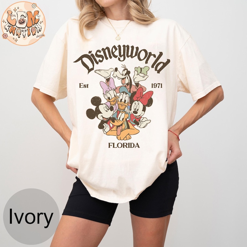 K&ouml;nnte beinhalten: Ein wei&szlig;es T-Shirt mit einer Grafik im Vintage-Stil von Mickey Mouse, Minnie Mouse, Donald Duck, Goofy und Pluto. Die Grafik enth&auml;lt den Text "Disneyworld Est 1971 Florida".