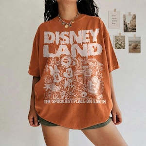 May include: Orange t-shirt featuring a distressed "DISNEY LAND" graphic and a spooky cartoon scene with Mickey and Minnie Mouse. The text "THE SPOOKIEST PLACE ON EARTH" is printed below. The shirt has an oversized fit.