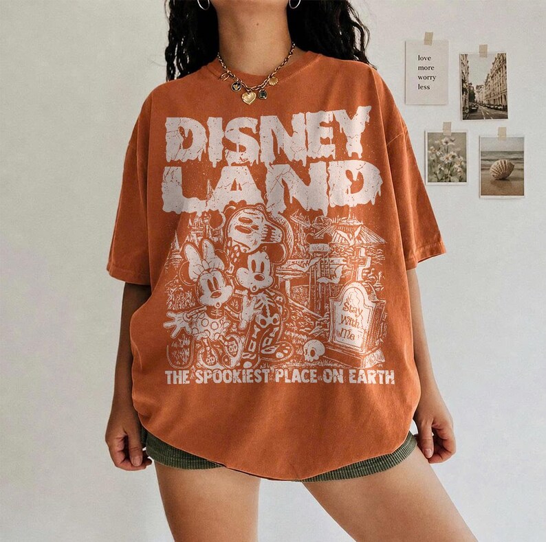 May include: Orange t-shirt featuring a distressed "DISNEY LAND" graphic and a spooky cartoon scene with Mickey and Minnie Mouse. The text "THE SPOOKIEST PLACE ON EARTH" is printed below. The shirt has an oversized fit.