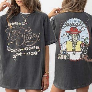May include: A charcoal gray t-shirt featuring a front graphic with rope-style text reading "It's a Toy Story baby just yes say" and a back graphic showing a cowgirl with a braid, a red hat, a cow-print boot, and blocks labeled "13 TS" with "Life of a Cowgirl" text.