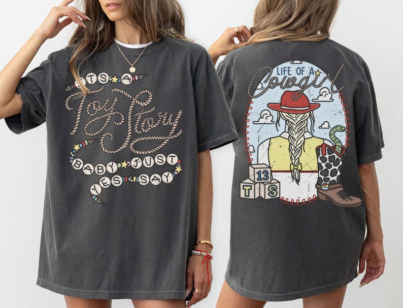 May include: A charcoal gray t-shirt featuring a front graphic with rope-style text reading "It's a Toy Story baby just yes say" and a back graphic showing a cowgirl with a braid, a red hat, a cow-print boot, and blocks labeled "13 TS" with "Life of a Cowgirl" text.
