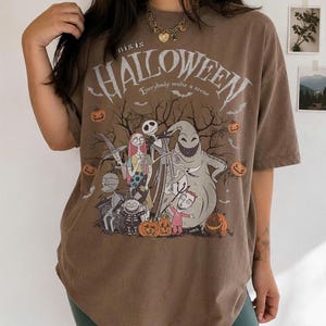 Pu&ograve; includere: T-shirt marrone con grafica a tema Halloween con personaggi e testo. Il design include Jack Skellington, Sally e Oogie Boogie, con le parole "this is HALLOWEEN" e "Everybody make a scene". Sono incluse anche zucche e pipistrelli.