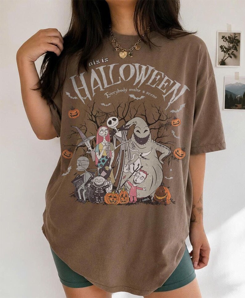 Pu&ograve; includere: T-shirt marrone con grafica a tema Halloween con personaggi e testo. Il design include Jack Skellington, Sally e Oogie Boogie, con le parole "this is HALLOWEEN" e "Everybody make a scene". Sono incluse anche zucche e pipistrelli.