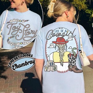 May include: A light blue short-sleeve t-shirt featuring a graphic on the back of a person with a braided ponytail wearing a red cowboy hat, a yellow vest, and a cow-print boot with a snake, plus three blocks labeled 13, T, and S.