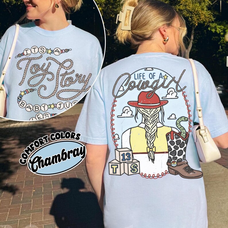 May include: A light blue short-sleeve t-shirt featuring a graphic on the back of a person with a braided ponytail wearing a red cowboy hat, a yellow vest, and a cow-print boot with a snake, plus three blocks labeled 13, T, and S.