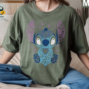 May include: A green t-shirt with a blue and purple floral Stitch design. The design features Stitch's signature big eyes and a floral pattern.