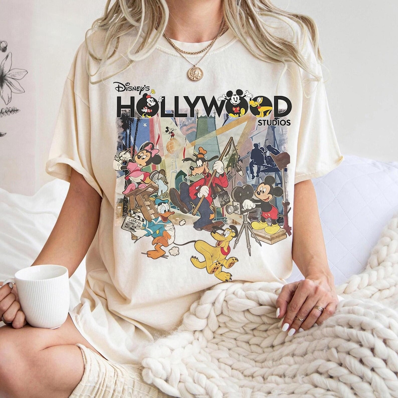 May include: Cream-coloured t-shirt featuring a graphic of Disney characters, including Mickey Mouse, Donald Duck, and Goofy, in a Hollywood Studios theme. The shirt has the text "Disney's Hollywood Studios" in a vintage style.